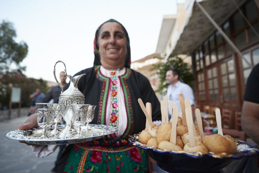 woman dressed with folklore cloths smiling and holding plateau with sweets and glasses walking at monastery of Ai Giannis area, in Lakki, Greece that commemorate festival of her birth