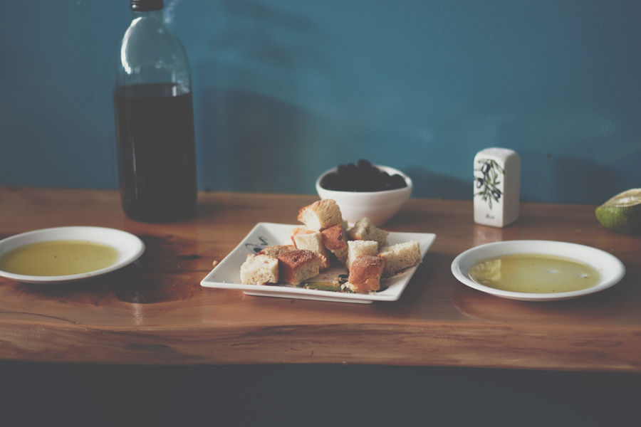 Against the backdrop of a serene blue wall, a wooden table boasts two plates filled with luscious olive oil, accompanied by a bottle of the same. A plate of bread takes center stage, while a small bowl of olives adds a flavorful touch to the Mediterranean tableau.