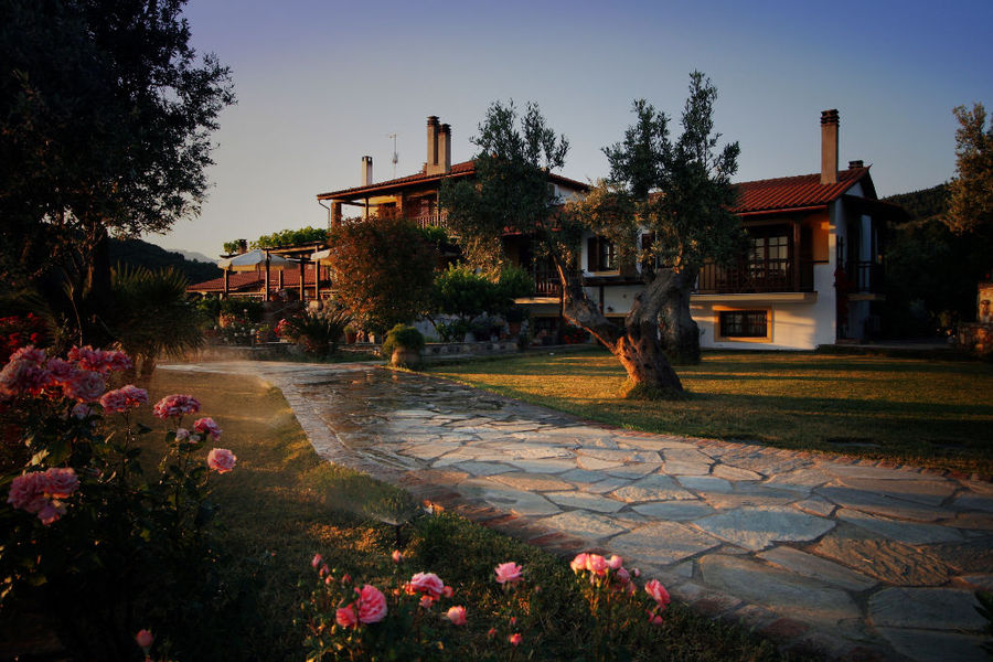 sunset at 'Vateri Guest House' complex buildings with stone alley in the front and trees and bushes of roses on the both sides