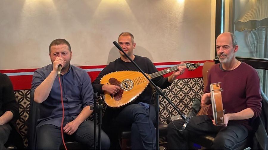 Three musicians perform live music at Ramon bar in Athens, playing instruments and singing into a microphone for the audience's entertainment.