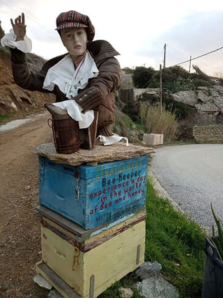 female doll mannequin with jacket and cap welcome on the dirt road at 'Melissavet' and two bee hives that says ' Bee-Keeper experience a day in the world of bee and honey'