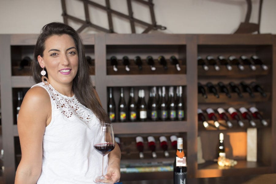 a smiling woman holding a glass of red wine in front of wooden shelves with bottles at 'Ancheon Winery'