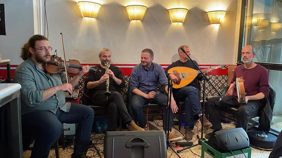 Five musicians perform live music at Ramon bar in Athens, playing various instruments and singing into a microphone for the audience's enjoyment.