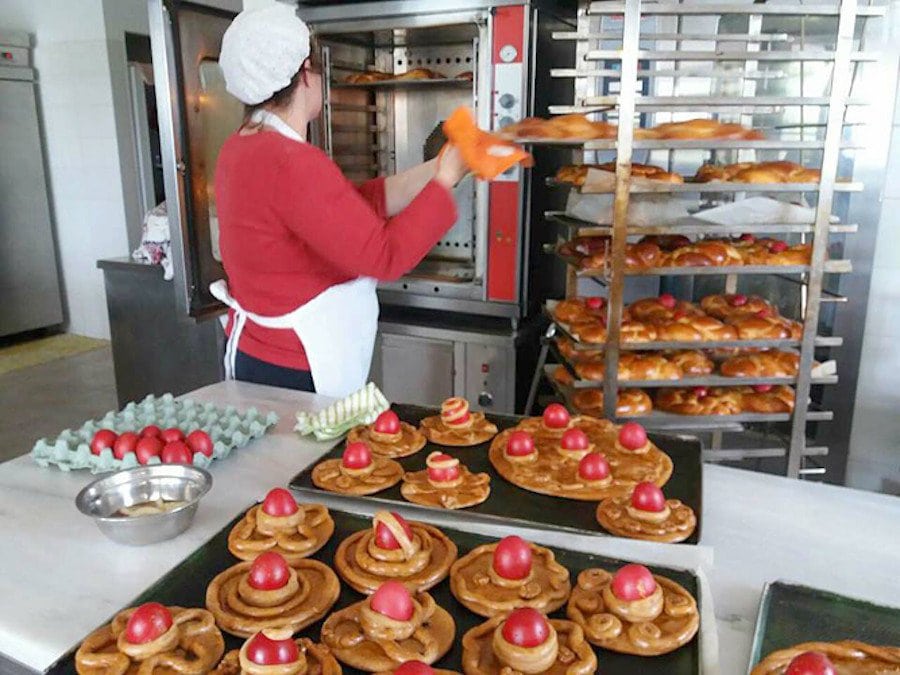 Woman putting bakery pan with Greek Easter cakes on metal trolley and surrounded by sweets with red eggs on the top