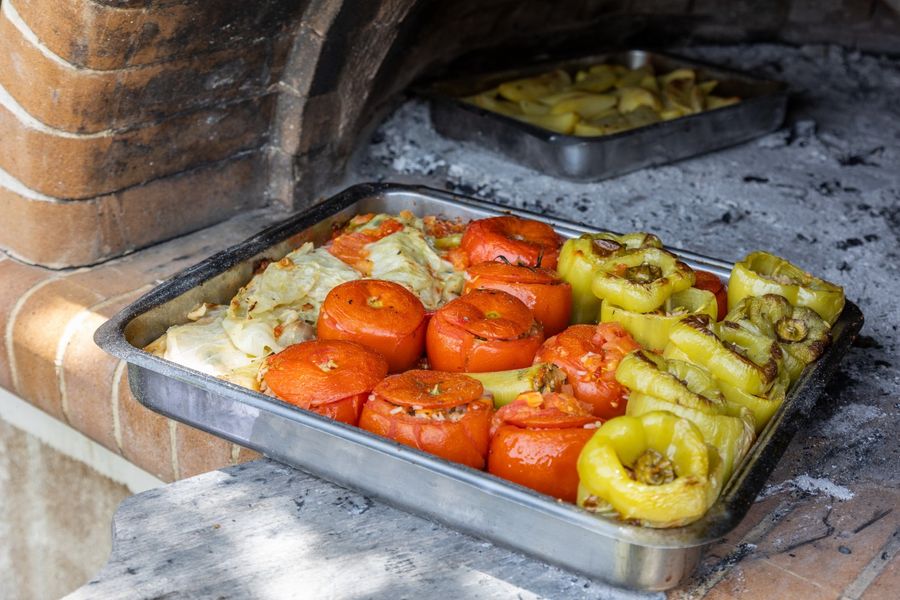 A baking pan filled with gemista, traditional Greek stuffed vegetables, rests on a wooden oven, the vibrant colors of the vegetables and aromatic herbs enticing the senses and promising a delicious Mediterranean feast.