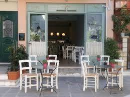 A picturesque Crete restaurant adorned with light blue wooden doors. Outside, white chairs and tables are elegantly arranged, inviting patrons to enjoy delightful cuisine in a serene and inviting ambiance.