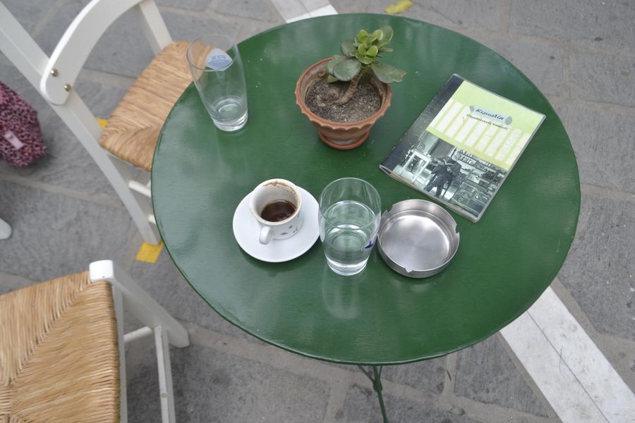 A green circular table with two glasses of water, a white cup of Greek coffee, a book, an ashtray, and a pot with a small plant at its center, epitomizing the harmony of nature and tranquility in a single captivating image.