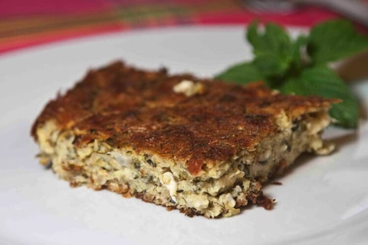 close-up of piece of ‘Kolokythopita’ means zucchini pie with green herbs in the background 