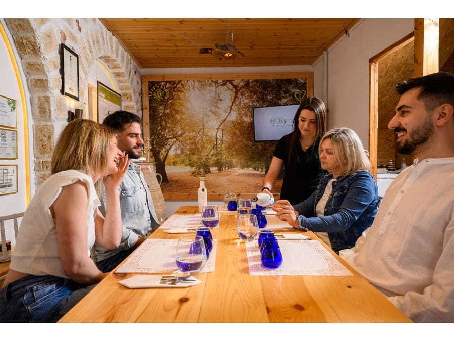 Four enthralled individuals gather around a meticulously set table for an olive oil tasting. A gracious woman pours golden oil into blue glasses, evoking a sense of anticipation and culinary delight.