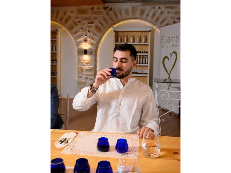 A man savors olive oil from a blue glass, captivated by its flavors. Before him, a place mat displays two more blue glasses, a glass of water, and a neatly folded napkin with fork and knife.