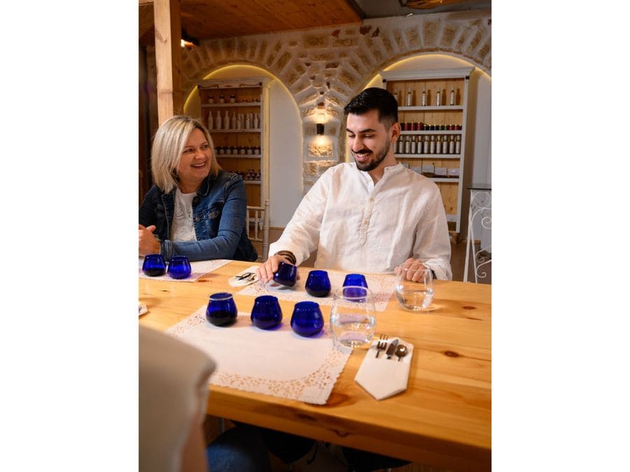 A man sits at an olive oil tasting table. Before him, a place mat displays three blue glasses and a glass of water, inviting a sensory exploration of flavors and aromas.