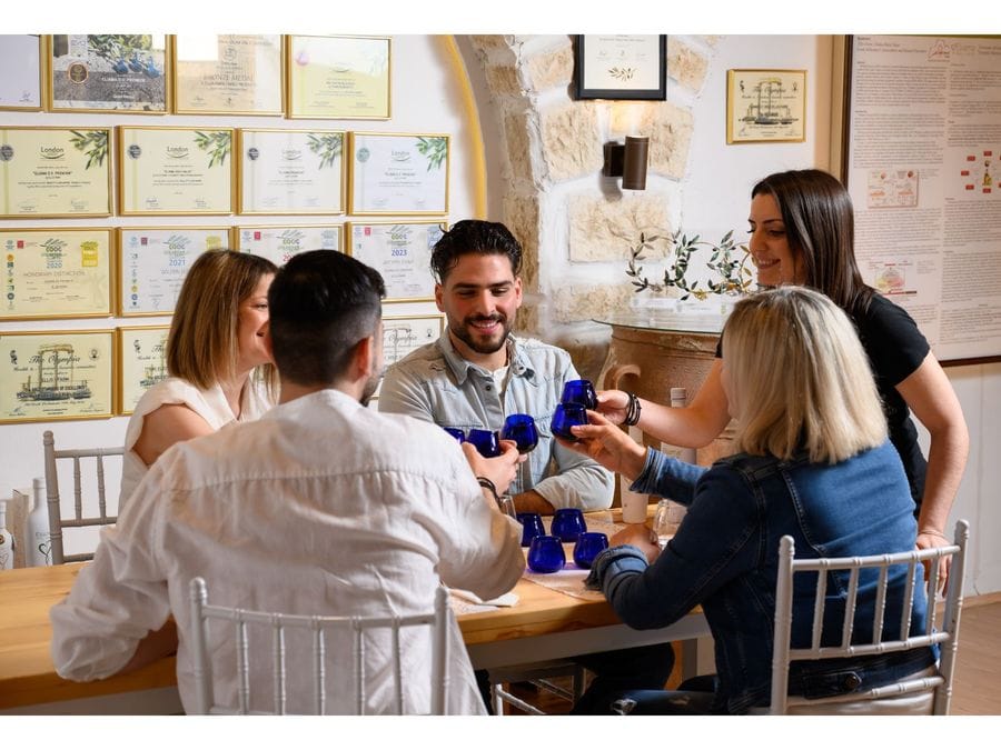 Five delighted individuals gather around a beautifully arranged table for an olive oil tasting. With glasses in hand, they joyfully clink them together, celebrating the shared experience of savoring this liquid treasure.