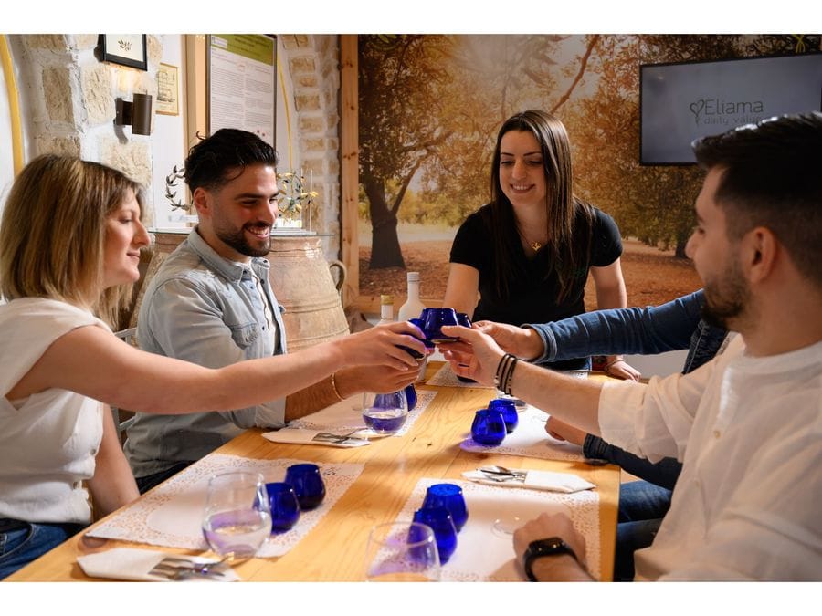 Five delighted individuals raise their blue glasses filled with olive oil, clinking them in unison. The shared moment exudes happiness and gratitude, symbolizing appreciation for the flavors and the companionship that the olive oil brings to their gathering.