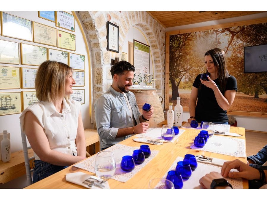 Four people gather around a table set for olive oil tasting. Place mats hold three blue glasses, a water glass, and utensils wrapped in a napkin. A standing woman passionately explains the art of olive oil tasting, setting the stage for a captivating and educational experience.