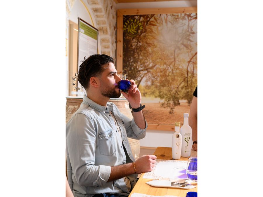 A man deeply engrossed in the olive oil tasting experience. With a blue glass in hand, he passionately smells and tastes the oil, radiating fascination and captivation. The table is meticulously set, inviting exploration and enjoyment.