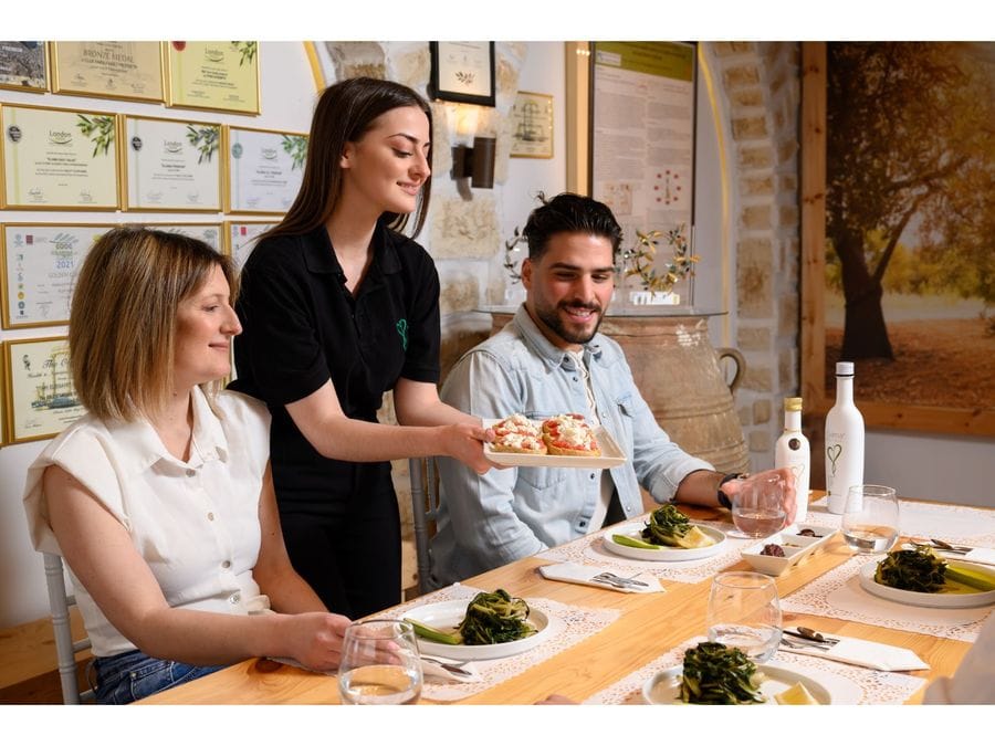 A gracious woman serves delectable delicacies to two seated individuals, complementing the olive oil tasting experience with carefully chosen treats, adding an exquisite touch to their gastronomic journey.