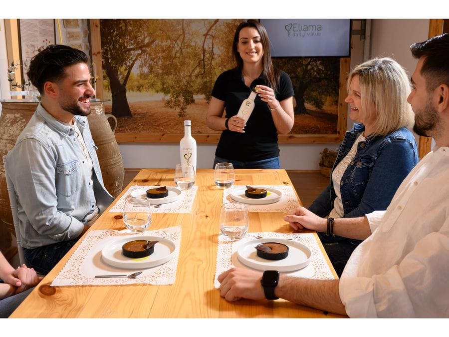 Four people gathered around a table for olive oil tasting. Sweet treats on plates, a bottle of water, and a standing woman holding olive oil. Pure joy fills the air.