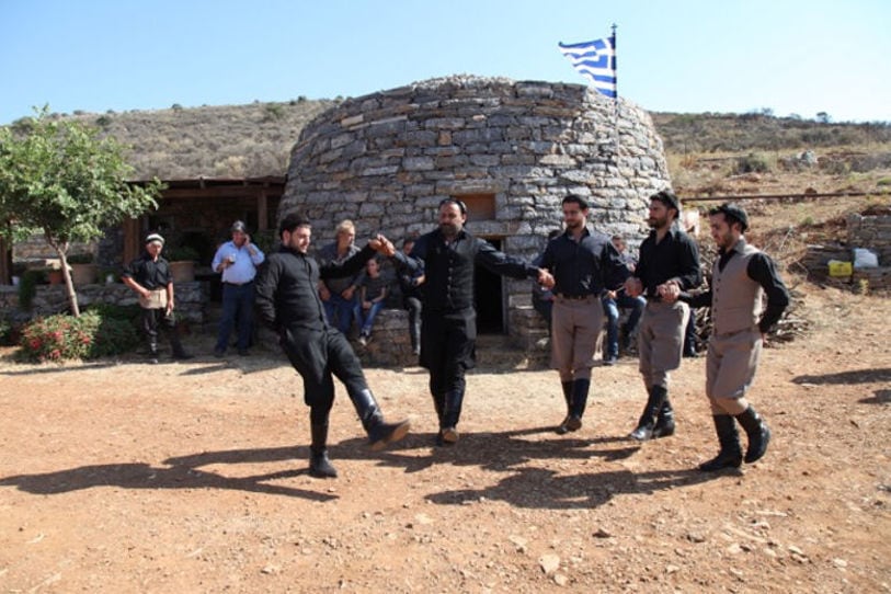 five men dancing traditional Cretan dance in front of small stone building with Greek flag at 'Galeni' farm