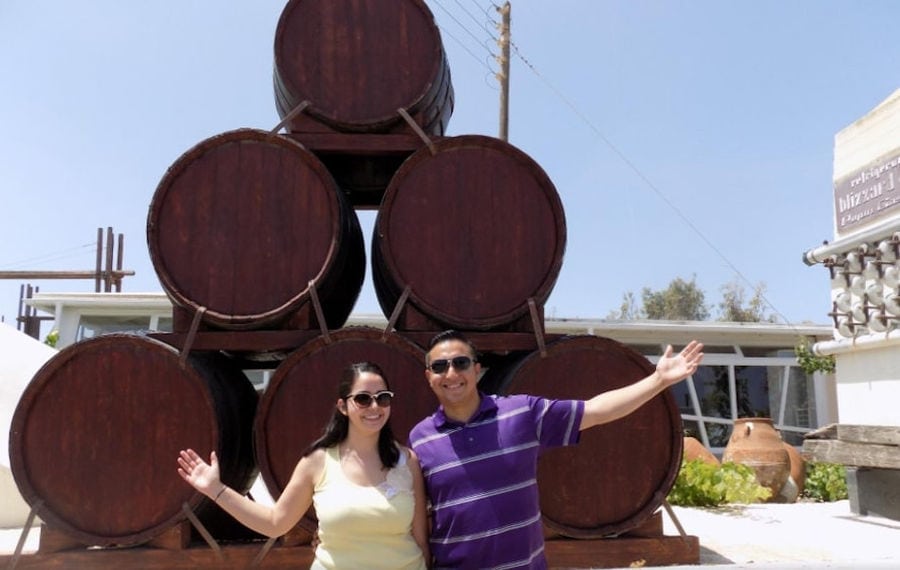 couple smiles happily at the camera in the background of wood wine barrels at 'Koutsoyannopoulos winery'