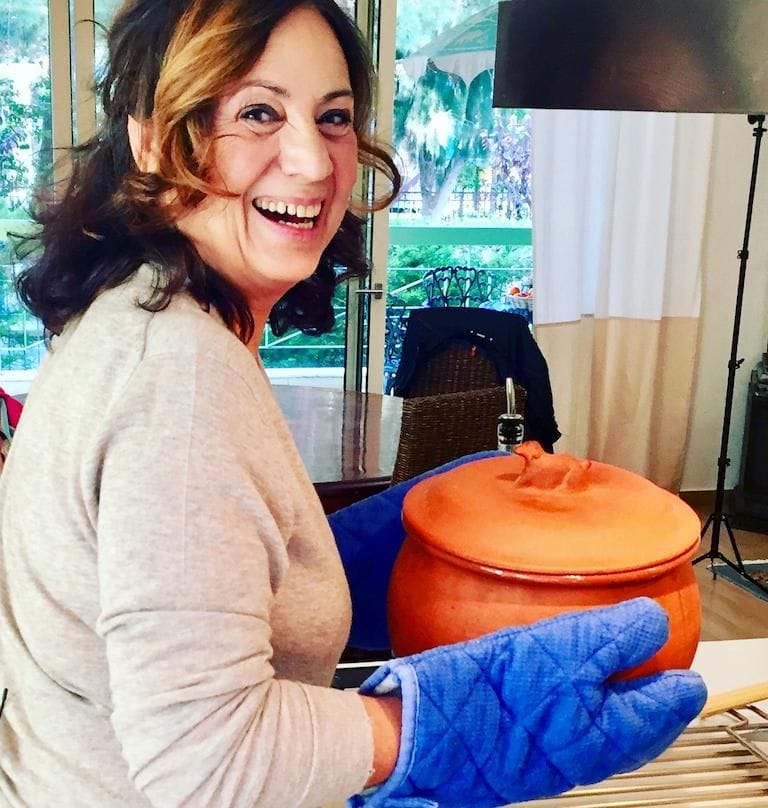 a woman holds a pot with hot food and smiles at the camera from Greek Cooking Class from Two Minutes Angie