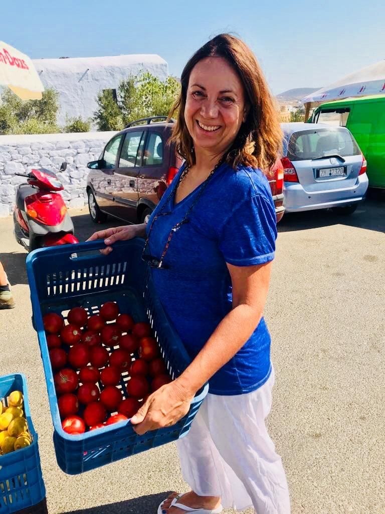 a woman smiles at the camera and holds a crate of tomatoes from Greek Cooking Class from Two Minutes Angie