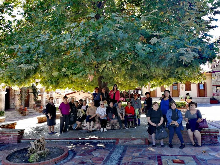 a group of tourists in the shade of the tree at 'Agios Antonios Women’s Agri Cooperative' stone building and watching at the camera