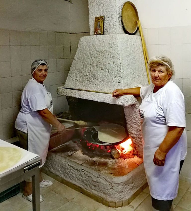 two woman baking pita bread in the fireplace and watching at the camera at 'Agios Antonios Women’s Agri Cooperative'