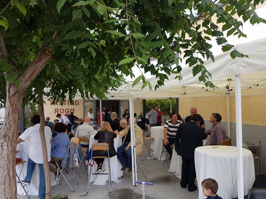 tourists tasting wines at the tables outside of 'Alexakis Winery' building that says 'TASTING ROOM' on the wall