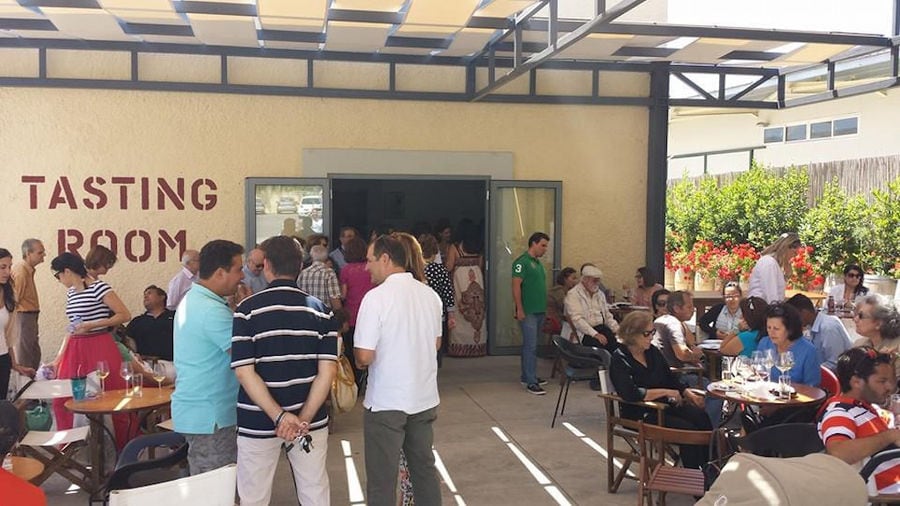 tourists tasting wines at the tables outside of 'Alexakis Winery' building that says 'TASTING ROOM' on the wall