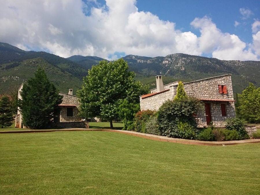 complex of Amfikaia stone buildings surrounded by green lawn and trees and mountains on the background