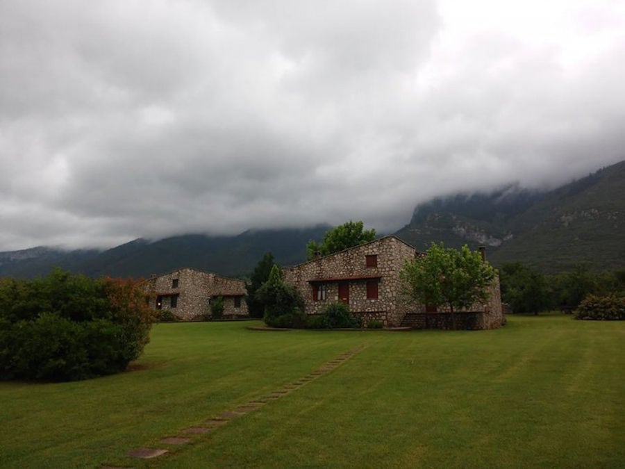 complex of Amfikaia stone buildings surrounded by green lawn and trees and mountains on the background