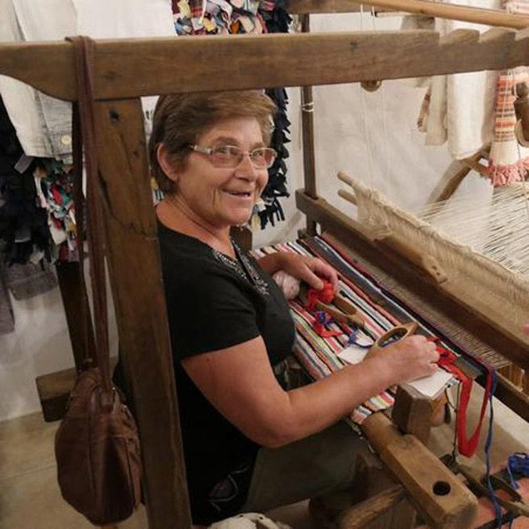 old smiling woman sitting on traditional loom, making carpets at Aristaios'