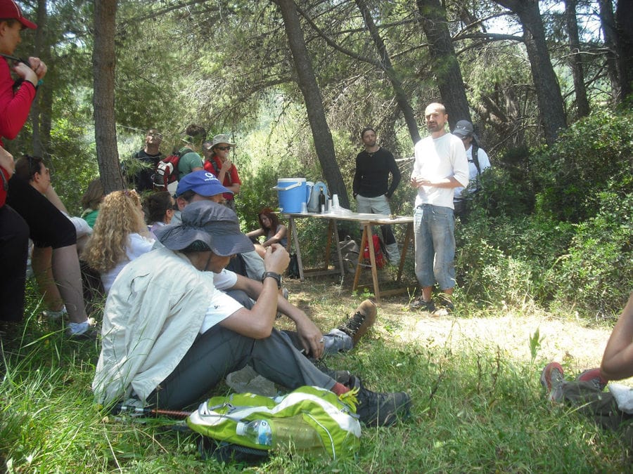 a group of tourists listening to a guide from 'Astarti' surrounded by trees and giving cooking lessons