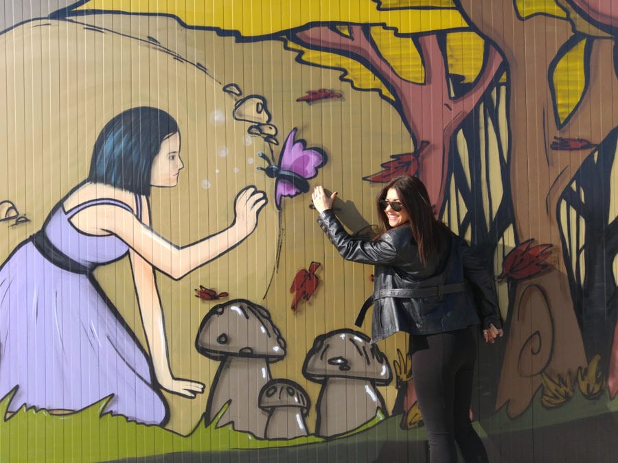 a woman showing to the camera a 'Dirfis' painted wall represents girl and butterfly surrounded by mushroom