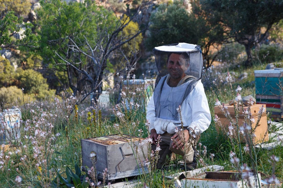 beekeeper smiling at the camera and holding a bee hive smoker at 'Eumelon' surrounded by white flowers and grass