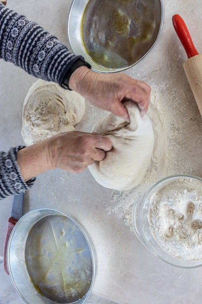 Amidst pans of olive oil and flour, a woman skillfully kneads dough, her hands moving with grace and purpose, embodying the artistry of breadmaking in a captivating culinary scene.