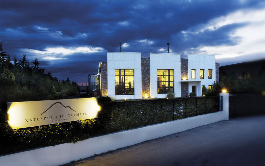 illuminated 'Katsaros Distillery' building by night with plants fence in the front