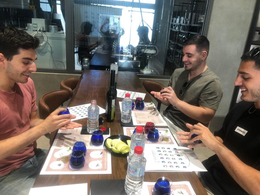 Three men gather around a table set for olive oil tasting. With blue glasses in hand, they savor the exquisite flavors. Each man has a personal place mat, complete with additional glasses, water bottles, and a central plate adorned with sliced green apples.