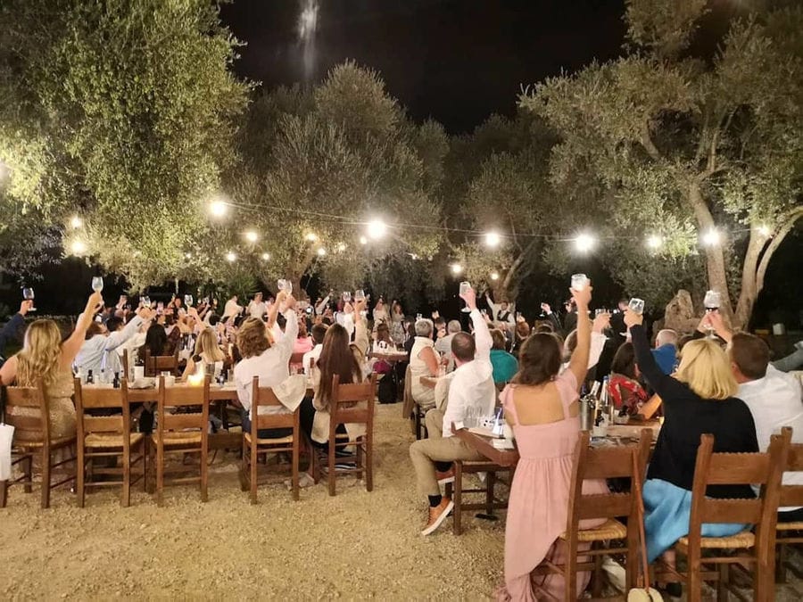 tourists sitting at the tables and raise their glasses of wine for 'cheers' at illuminated Manousakis Winery by night