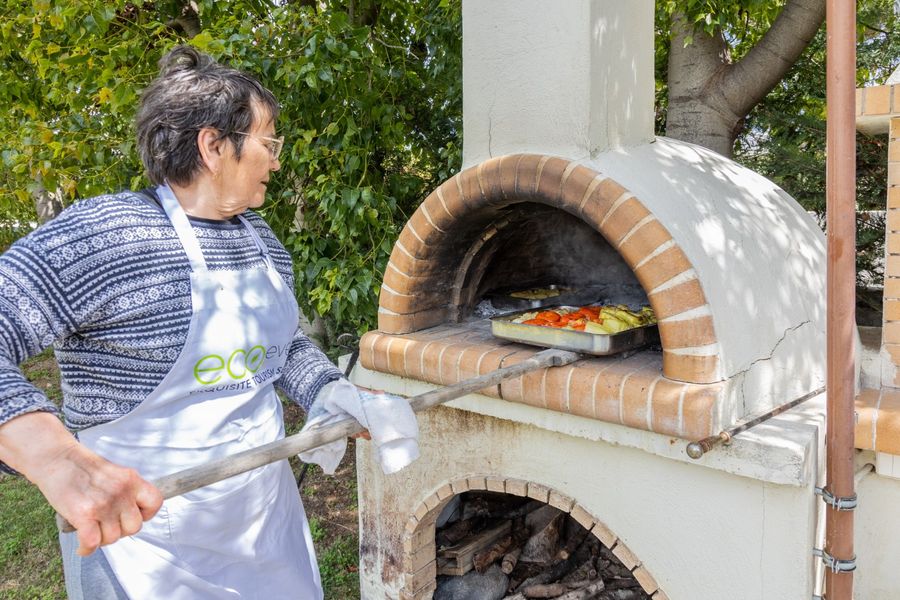 A woman gently slides a baking pan filled with gemista into a wooden oven, the vibrant colors of the stuffed vegetables sizzling and blending, as the oven's warmth promises to bring out their rich flavors and create a mouthwatering meal.