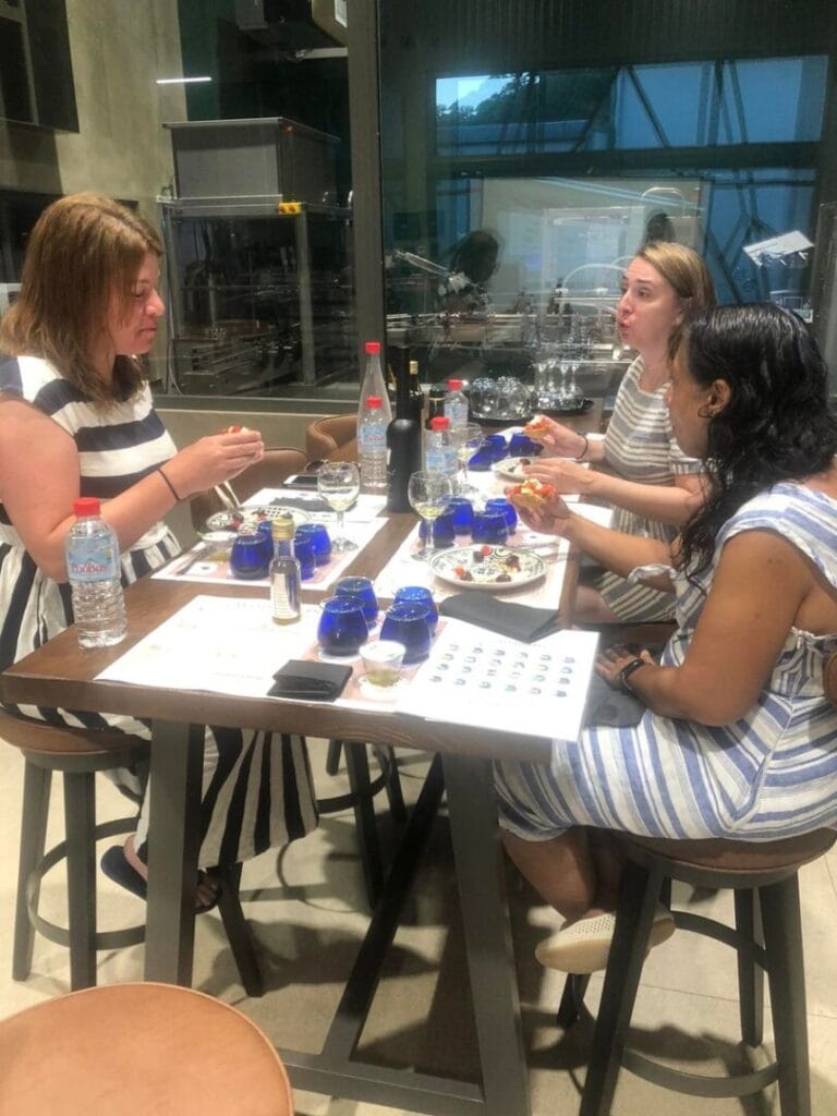Three delighted women gather around a beautifully set table for olive oil tasting. Their joyful expressions say it all as they indulge in ntakos, savoring the delightful flavors of olive oil.