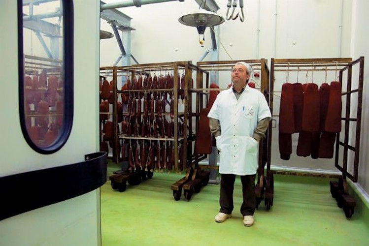 man in white robe standing in front of hangers with processed meat at 'Sary'