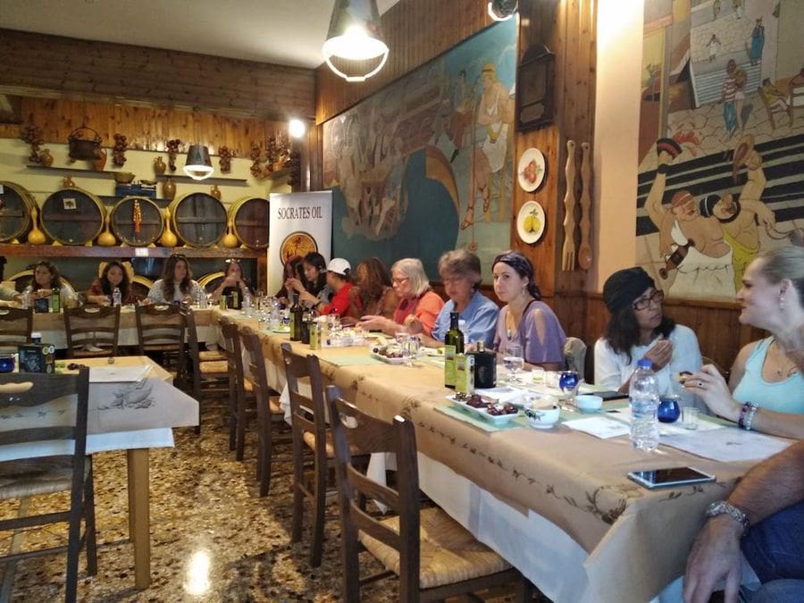 tourists sitting at the tables and tasting olive oil and olives in tasting room with a painted fresco on one wall and wooden barrels on another at Socrates Olive Oil