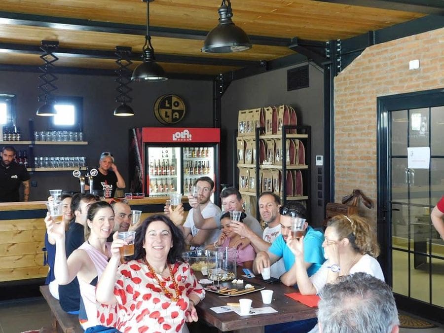 tourists smiling happily at the camera, sitting at the table and raise their glasses of beer for 'cheers' at Pineios Brewery tasting room