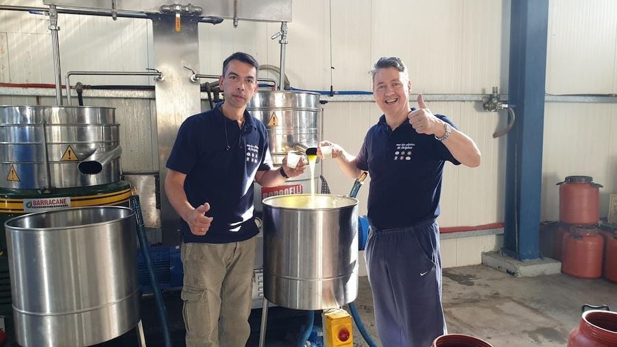 two men smiling at the camera and holding glasses with olive oil pointing with fingers ok at Mer des Oliviers de Delphes premises