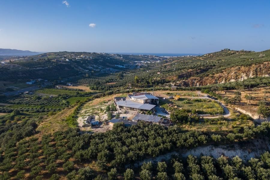 view from above of bee hives under the sunshades and a building of Kokkiadis honey farm on the left surrounded by trees, hills and a blue sky in the background