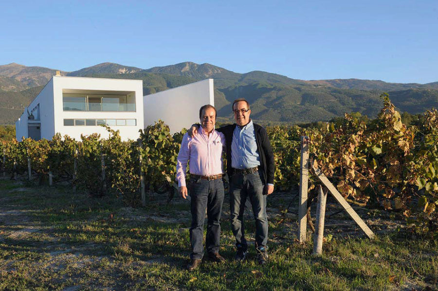 two men smiling happily at the camera in the background of vineyards and 'Voyatzis Estate' building