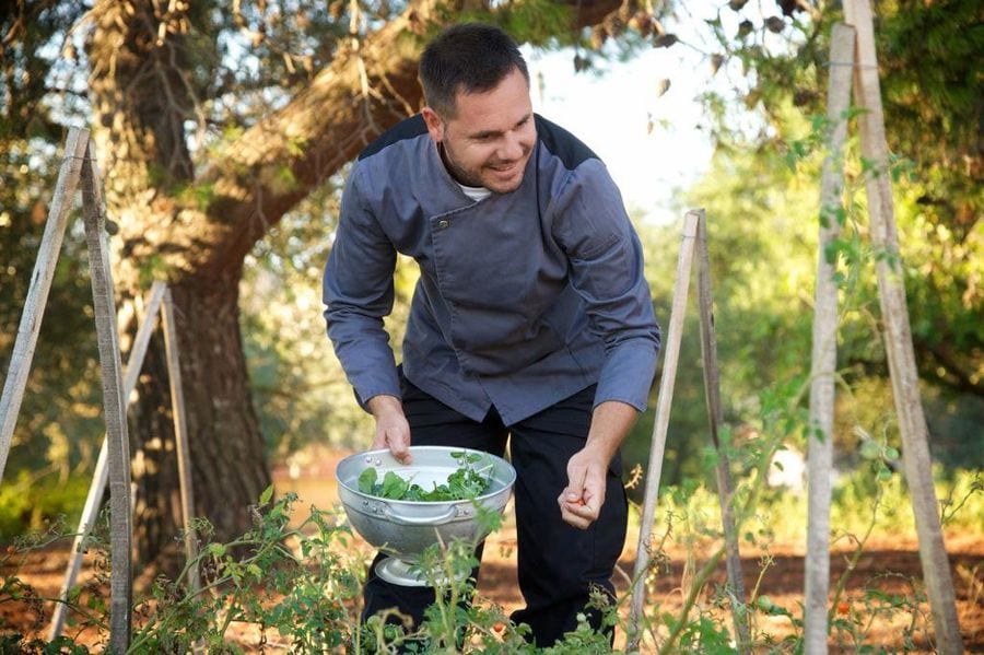 a smilling young man holding a small vase with green plants with trees in the background at En Kefallinia Organic Farm Restaurant garden